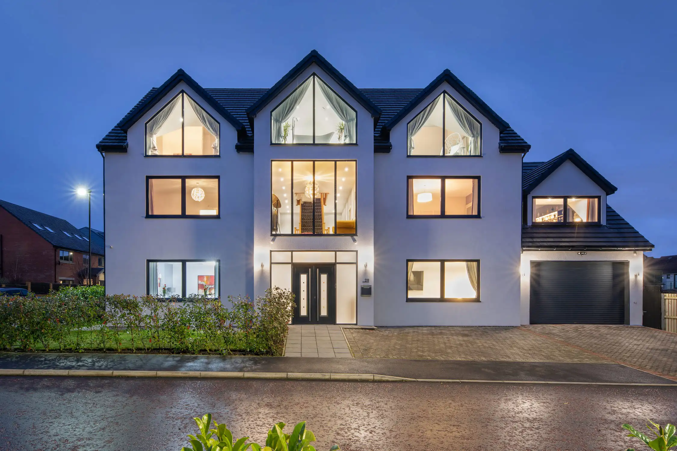 Large self build home designed by Acre Design Newcastle on Hazelwood Road, Great Park. White rendered home with a grey pitched roof. Photographed at night with warm glow from the inside. Open plan family home designed with large family bathrooms, dressing rooms and luxurious finishes.
