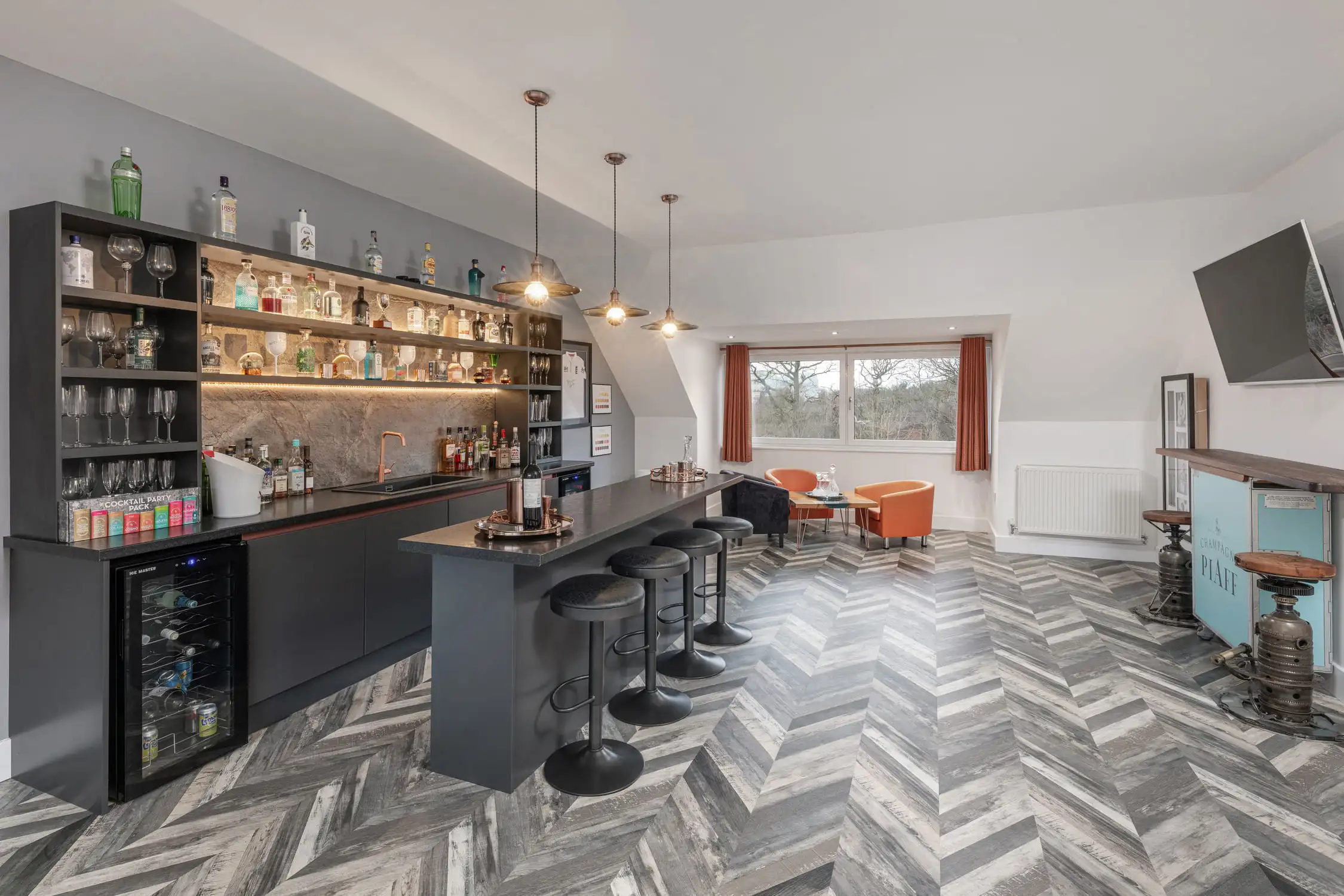 Hazelwood-Road-Great-Park-Gosforth-Newcastle-Self-Build-Home-Design-Acre-home-bar-private-luxury This home bar features a large island with bar stools, a fully stocked drinks shelf and beer taps. There are four bar stools and some tub chair seats too. The grey herringbone floor adds to the quirky feel, coupled with the pendant lights overhanging the bar area. Large self build home designed by Acre Design Newcastle on Hazelwood Road, Great Park.