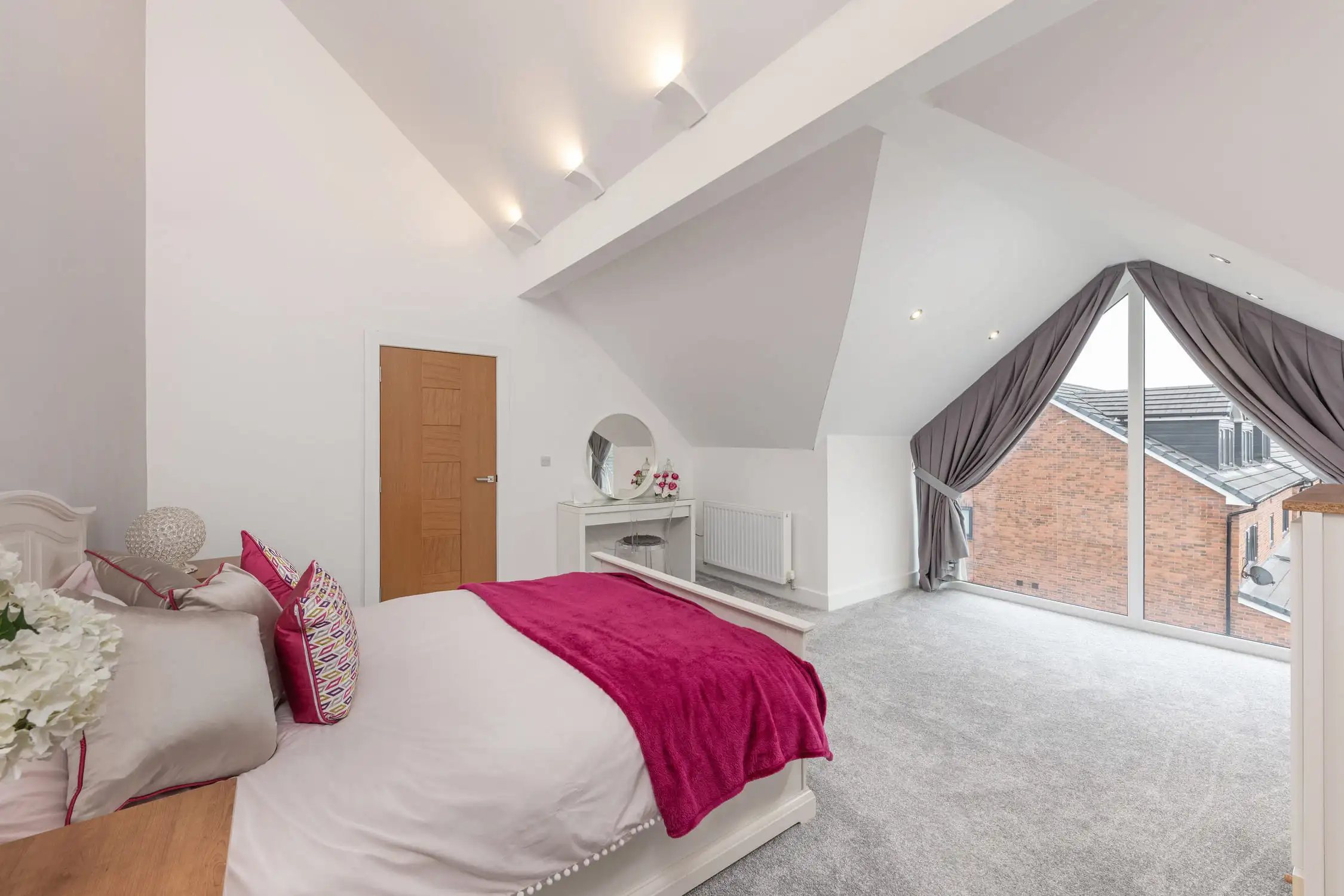 Hazelwood-Road-Great-Park-Gosforth-Newcastle-Self-Build-Home-Design-Acre-master-bedroom-suite-view-triangular-window-sash-curtain Large self build home designed by Acre Design Newcastle on Hazelwood Road, Great Park. A simple, light, open plan bedroom with a vaulted ceiling, looks out onto a large triangular window with a woodland view. White bedding with fuchsia pink bedspread and dark curtains. Fluffy Grey carpet and brown oak doors.