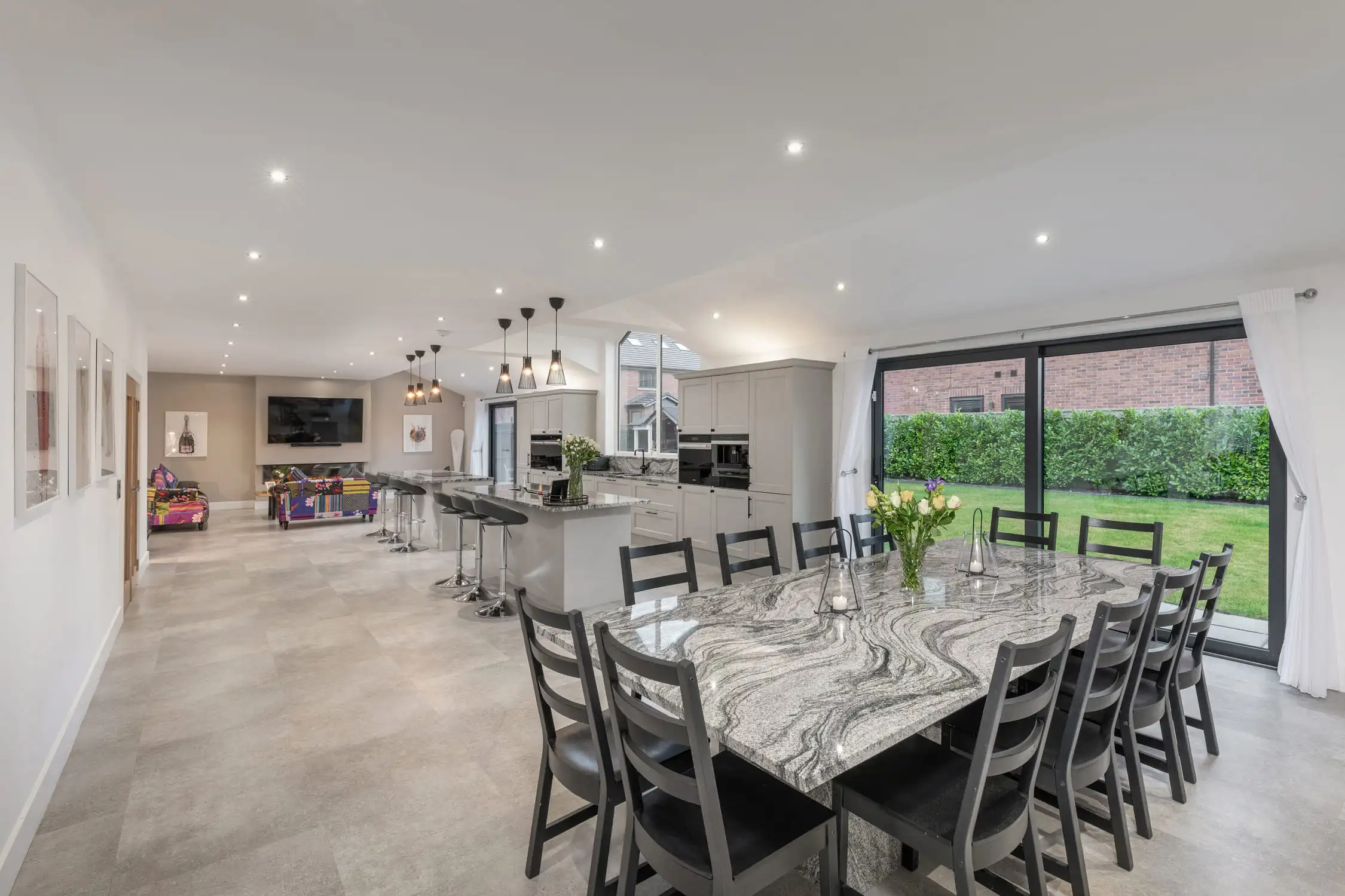 Hazelwood-Road-Great-Park-Gosforth-Newcastle-Self-Build-Home-Design-Acre-open-plan-kitchen-dining-living-luxury-dream Marble topped dining table to seat ten people frame the image of a large open plan kitchen living space. Plenty of natural light floods the room and it is also lit by pendant lighting and spotlights from above. There is a large flat screen tv on the wall and comfortable, patterned sofas to relax in. The kitchen island is large and welcoming. Large self build home designed by Acre Design Newcastle on Hazelwood Road, Great Park.