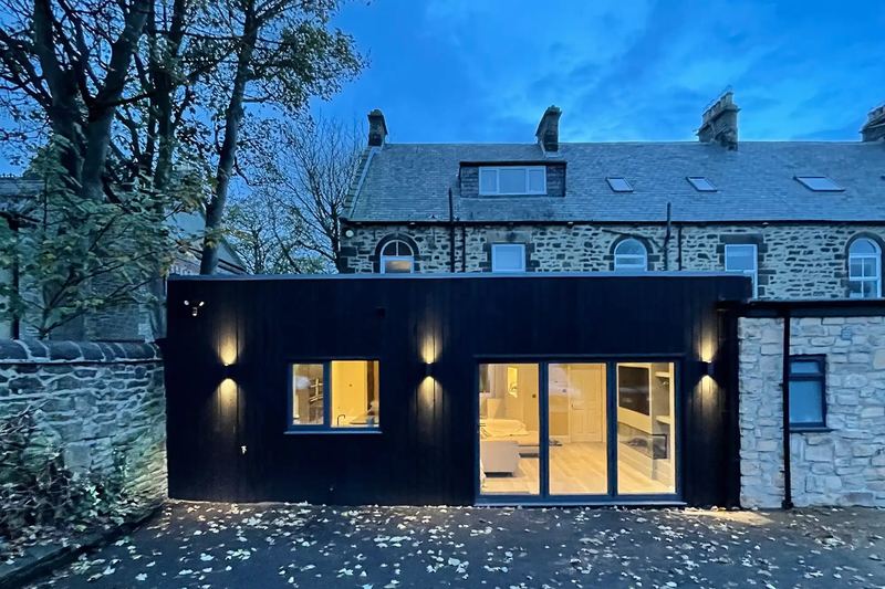 Completed rear house extension with charred larch cladding and full-height glazing, lit at dusk, on a stone house in Low Fell, Gateshead